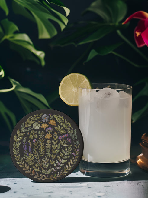 Circular ceramic coaster with black background featuring intricate botanical illustrations of various herbs, flowers and leaves in green, purple and gold colors with white ceramic edge and cork backing.